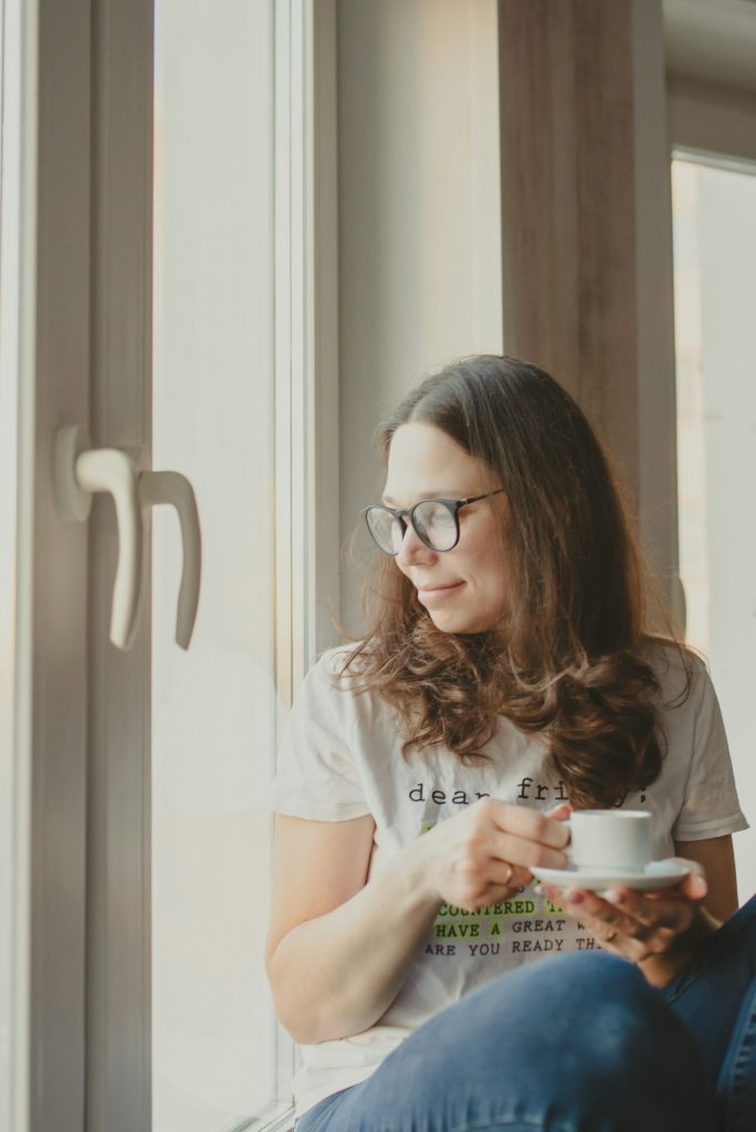 A young woman with glasses and loose hair drinks espresso with a smile and looks out the window.